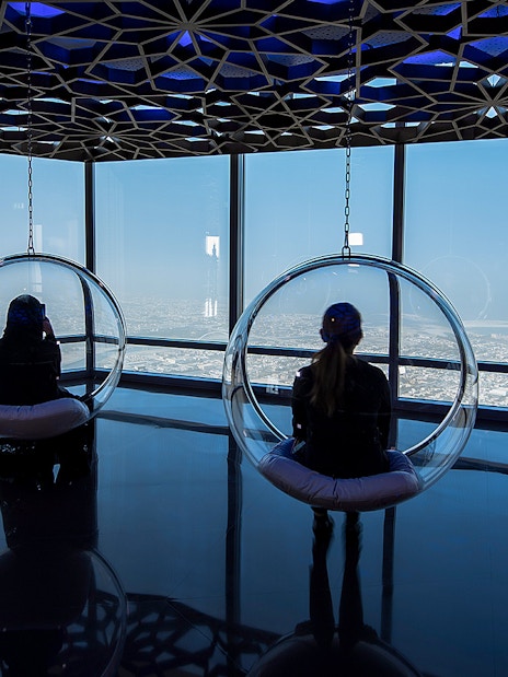 Visitors in glass chairs overlooking Dubai from Burj Khalifa observation deck.