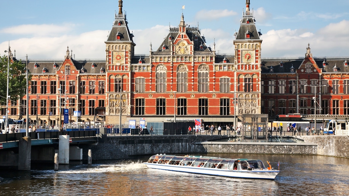 Cruise boat passing in front of Amsterdam Central Station under a bridge.