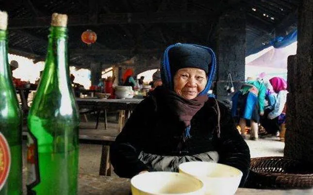 Elderly woman at Bac Ha Market with bottles and bowls on table, Vietnam.