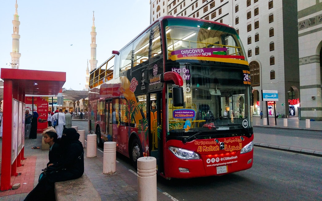 Red double-decker bus at Al Madinah Hop-On Hop-Off Tour stop with cityscape in background.