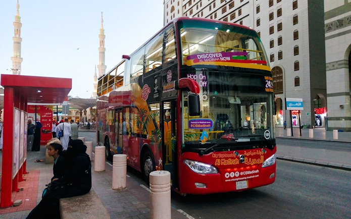 Red double-decker bus at Al Madinah Hop-On Hop-Off Tour stop with cityscape in background.