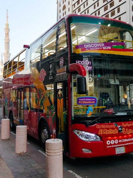 Red double-decker bus at Al Madinah Hop-On Hop-Off Tour stop with cityscape in background.