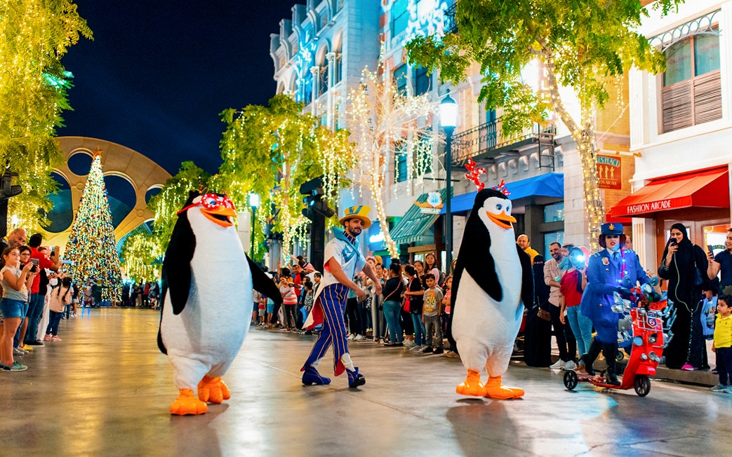 Penguin characters parade at MOTIONGATE™ Dubai with festive lights and crowd.