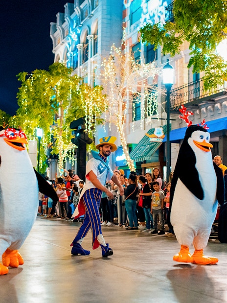Penguin characters parade at MOTIONGATE™ Dubai with festive lights and crowd.