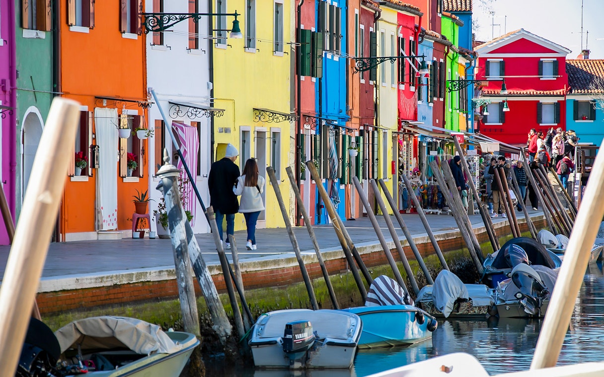 Colorful houses along a canal in Burano, Italy, with boats and tourists walking nearby.