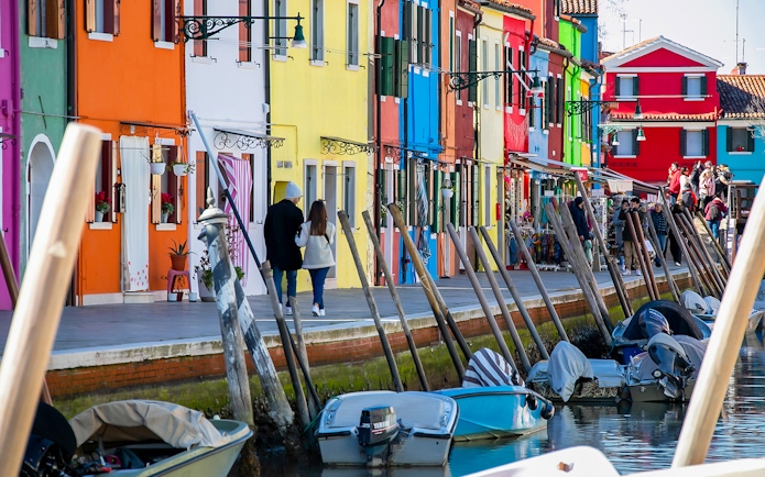 Colorful houses along a canal in Burano, Italy, with boats and tourists walking nearby.