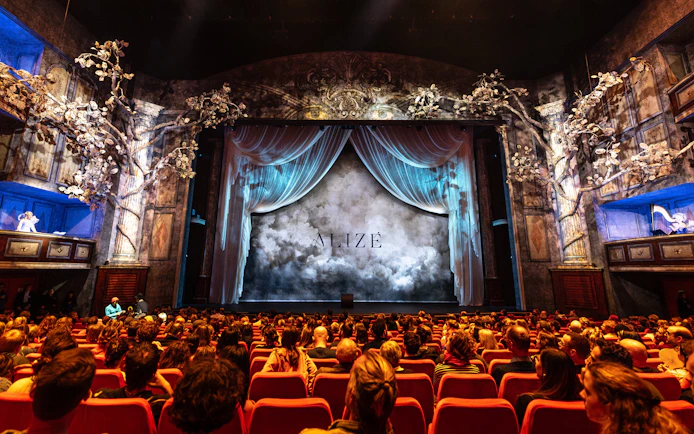 Audience seated in Alizé Theatre for Cirque du Soleil show, ornate stage with draped curtains.