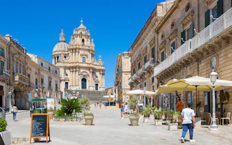 Baroque architecture in Ragusa, Italy, with people walking and dining outdoors.