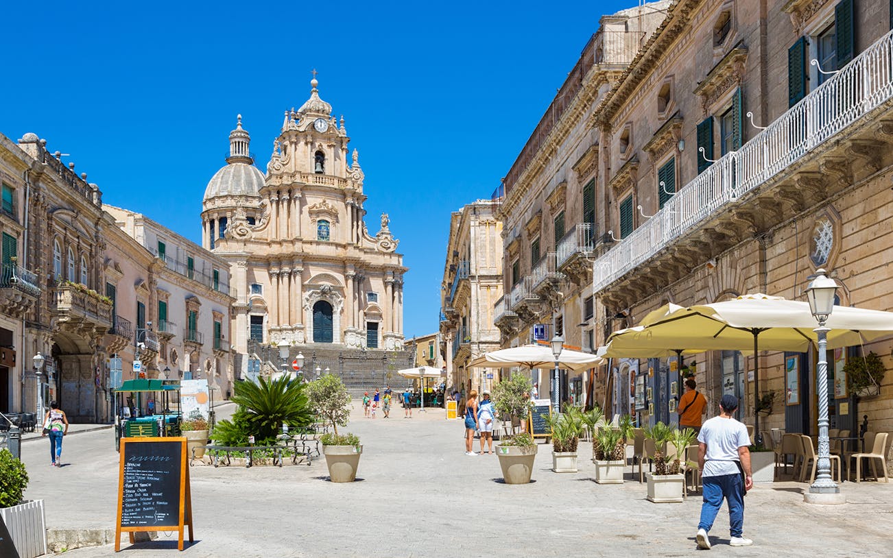 Baroque architecture in Ragusa, Italy, with people walking and dining outdoors.