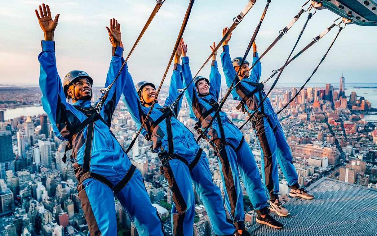 People in harnesses leaning over a skyscraper edge with New York City skyline in the background.