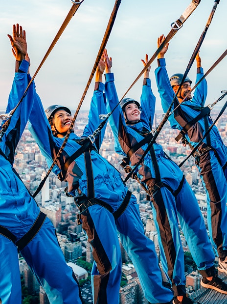 People in harnesses leaning over a skyscraper edge with New York City skyline in the background.
