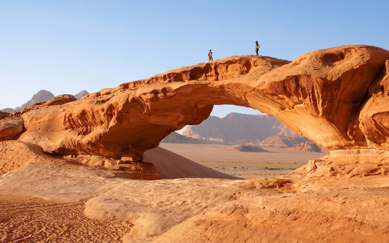 Natural rock arch in Wadi Rum desert with two people standing on top.