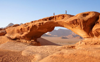 Natural rock arch in Wadi Rum desert with two people standing on top.