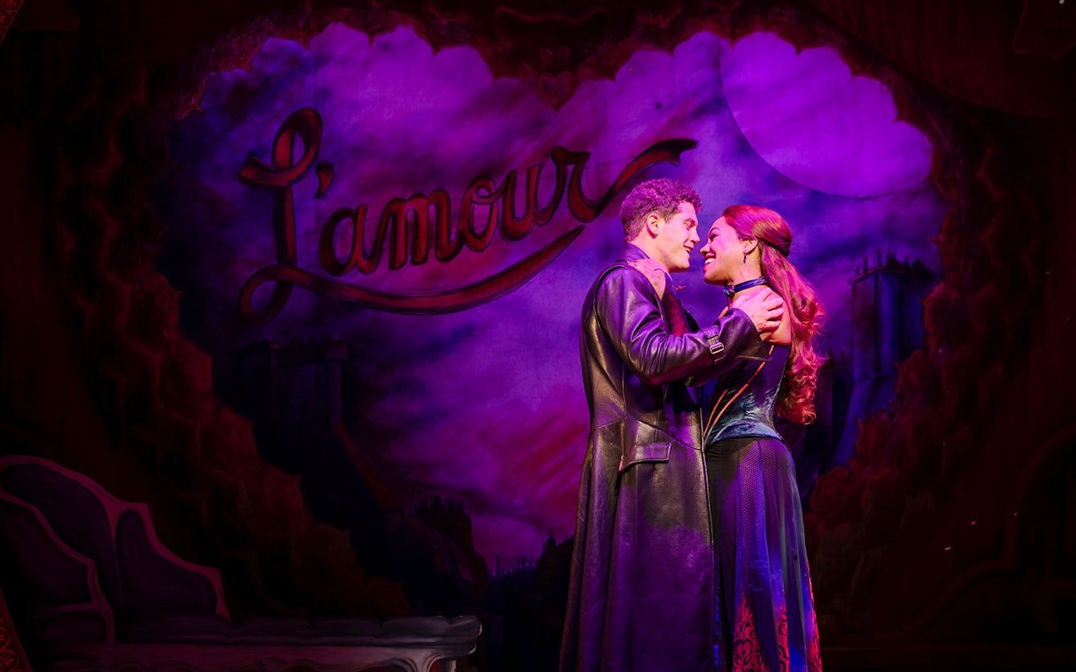 Performers embrace on stage during Moulin Rouge! The Musical in West End, with "L'amour" backdrop.