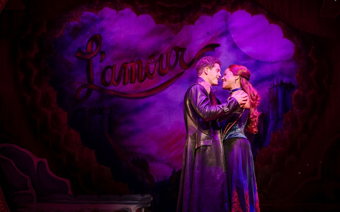 Performers embrace on stage during Moulin Rouge! The Musical in West End, with "L'amour" backdrop.