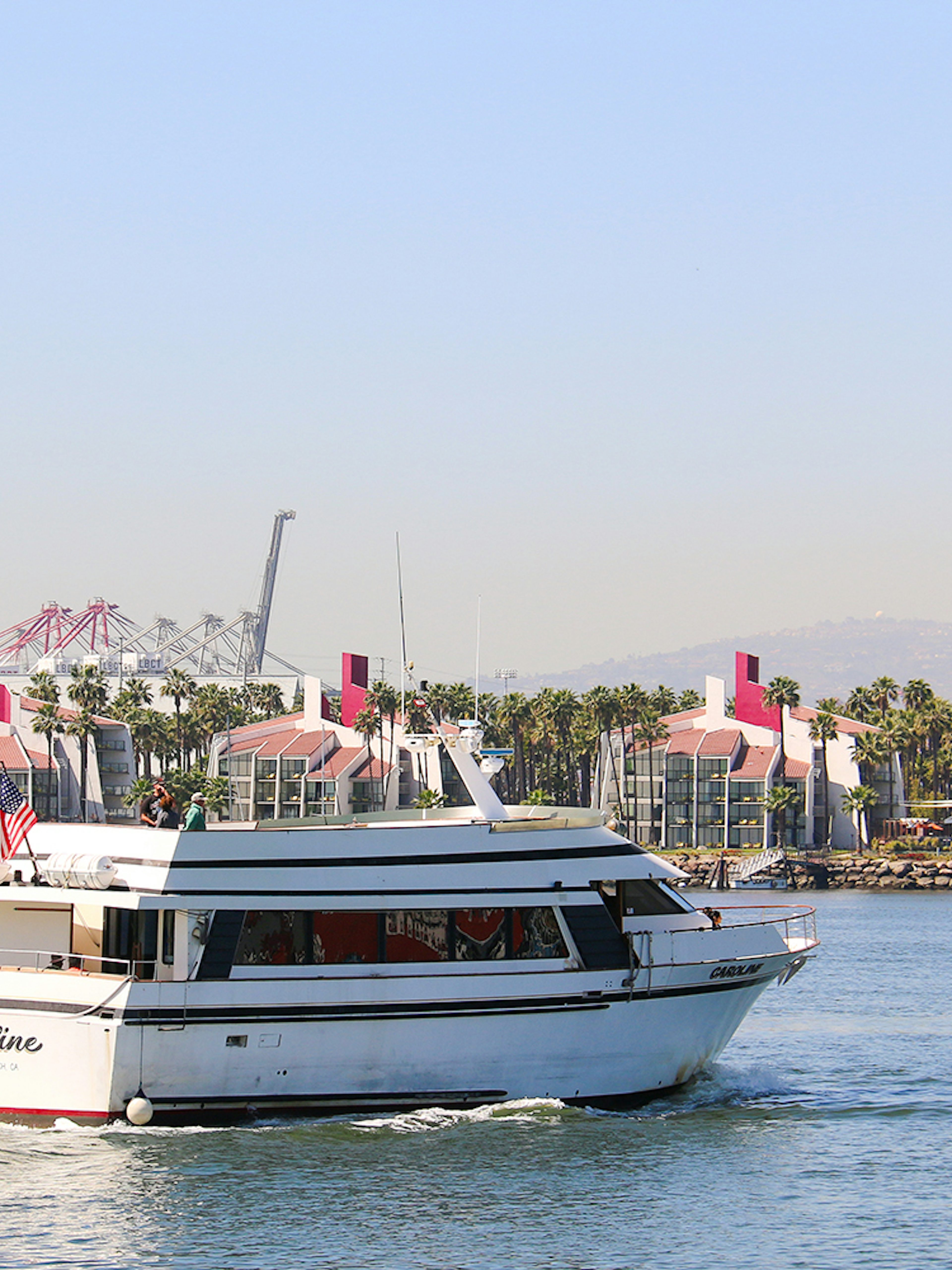 Cruise boat sailing in Long Beach, California with port cranes and palm trees in the background.