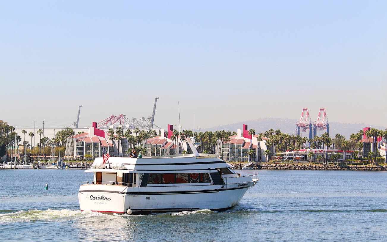 Cruise boat sailing in Long Beach, California with port cranes and palm trees in the background.