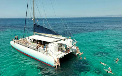 Catamaran with passengers on turquoise ocean, people swimming nearby under clear blue sky.