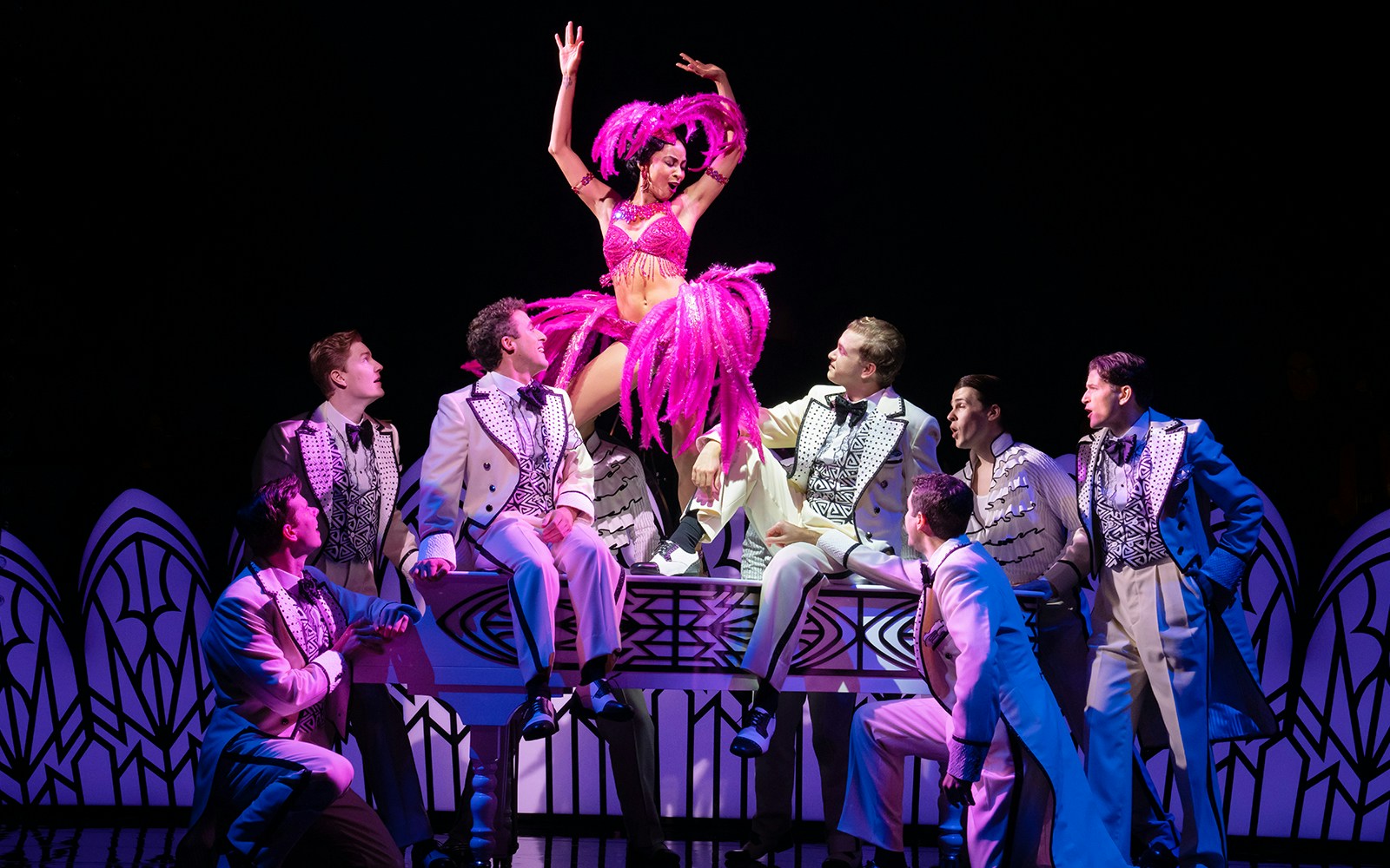 Performer in pink costume dancing on piano surrounded by men in suits, scene from Harmony: A New Musical.
