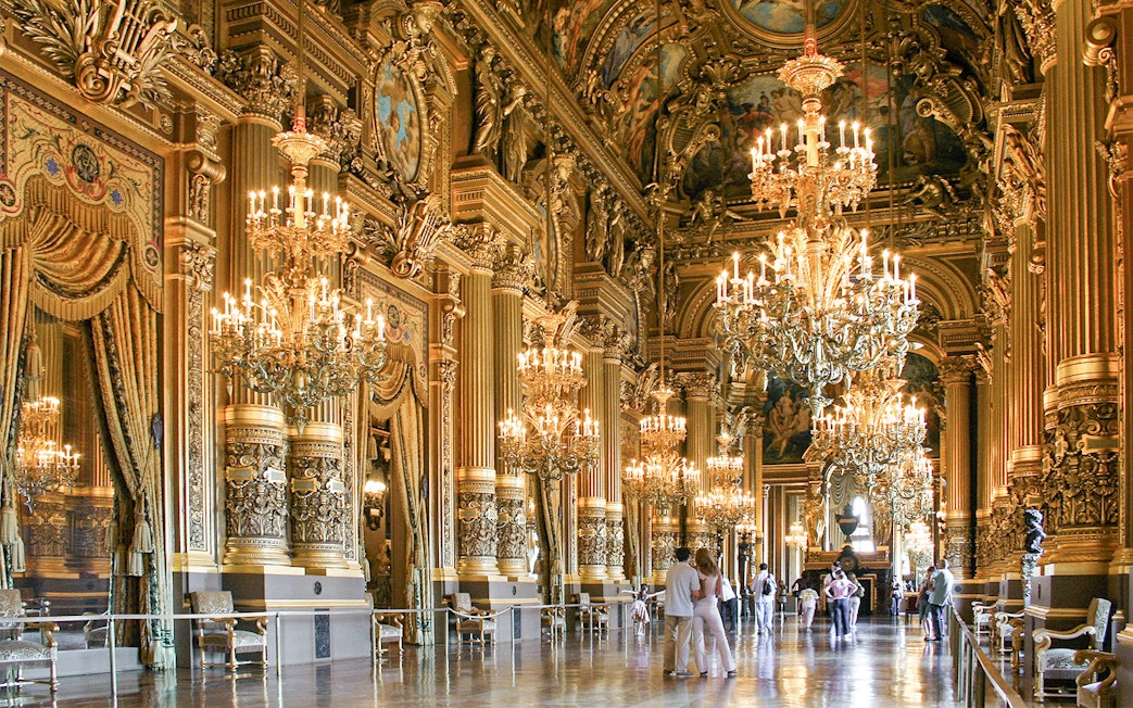 Visitors inside Opéra Garnier, Paris, admiring ornate chandeliers and gilded architecture.