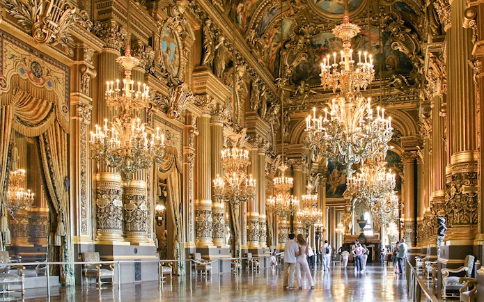 Visitors inside Opéra Garnier, Paris, admiring ornate chandeliers and gilded architecture.