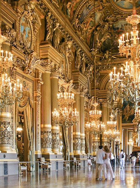 Visitors inside Opéra Garnier, Paris, admiring ornate chandeliers and gilded architecture.