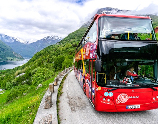 Red double-decker bus on Geiranger Hop-on Hop-off tour with fjord and mountains in background.