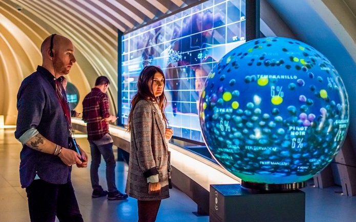 Tourists at Cité du Vin museum in Bordeaux using audio guides near interactive wine exhibit.
