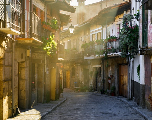 Narrow cobblestone street in Cuevas del Valle, Avila, with rustic buildings at sunset.