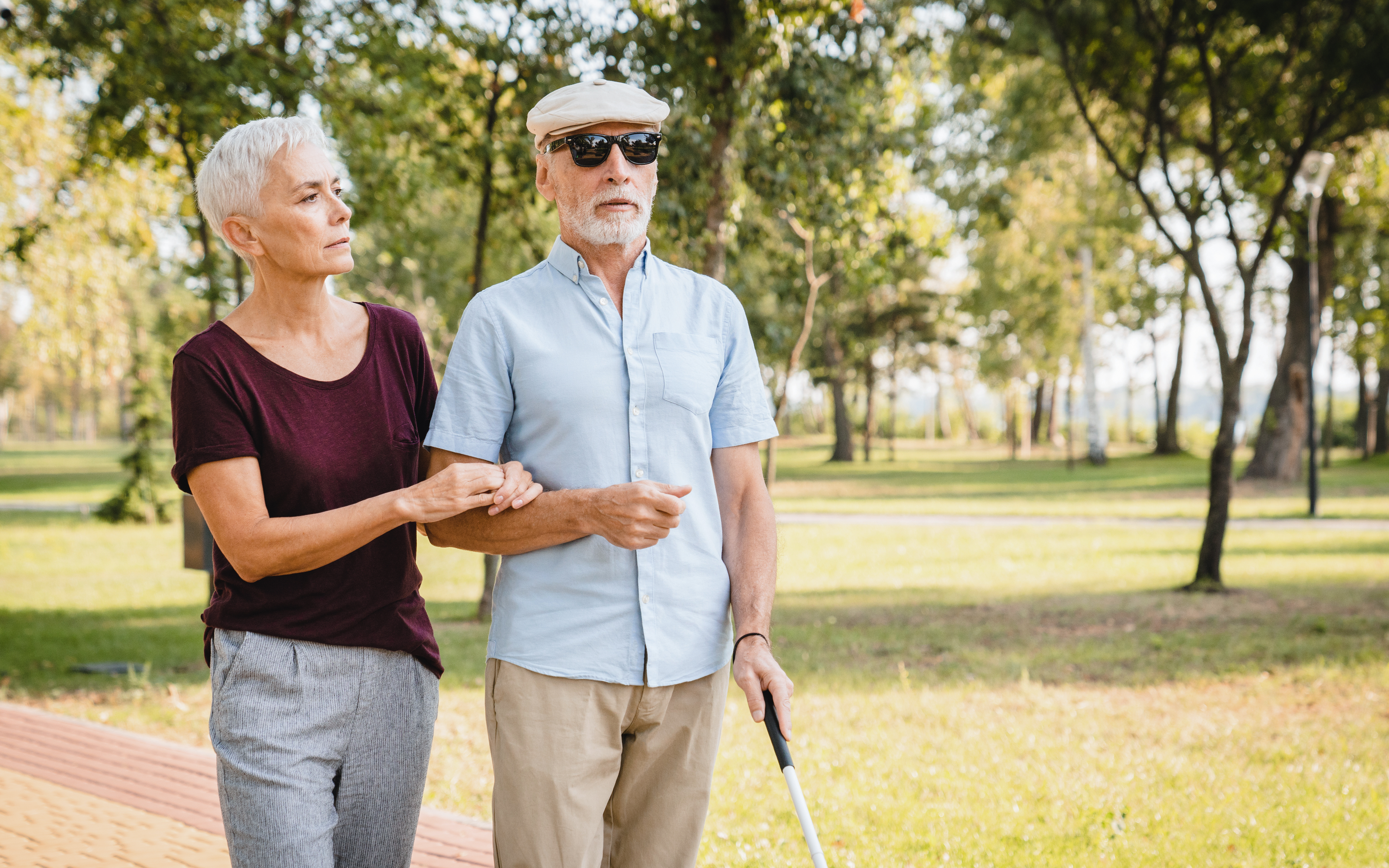 Elderly woman guiding visually impaired man with cane in a park.