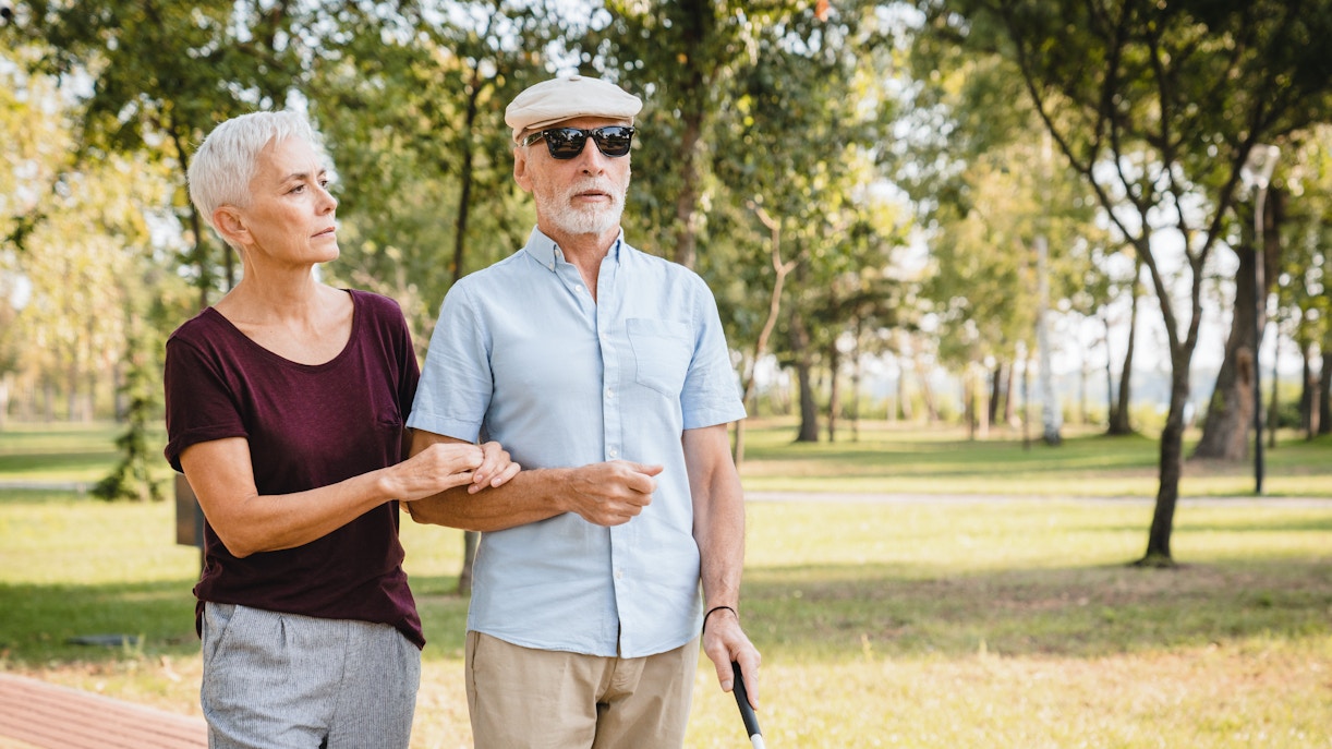 Elderly woman guiding visually impaired man with cane in a park.