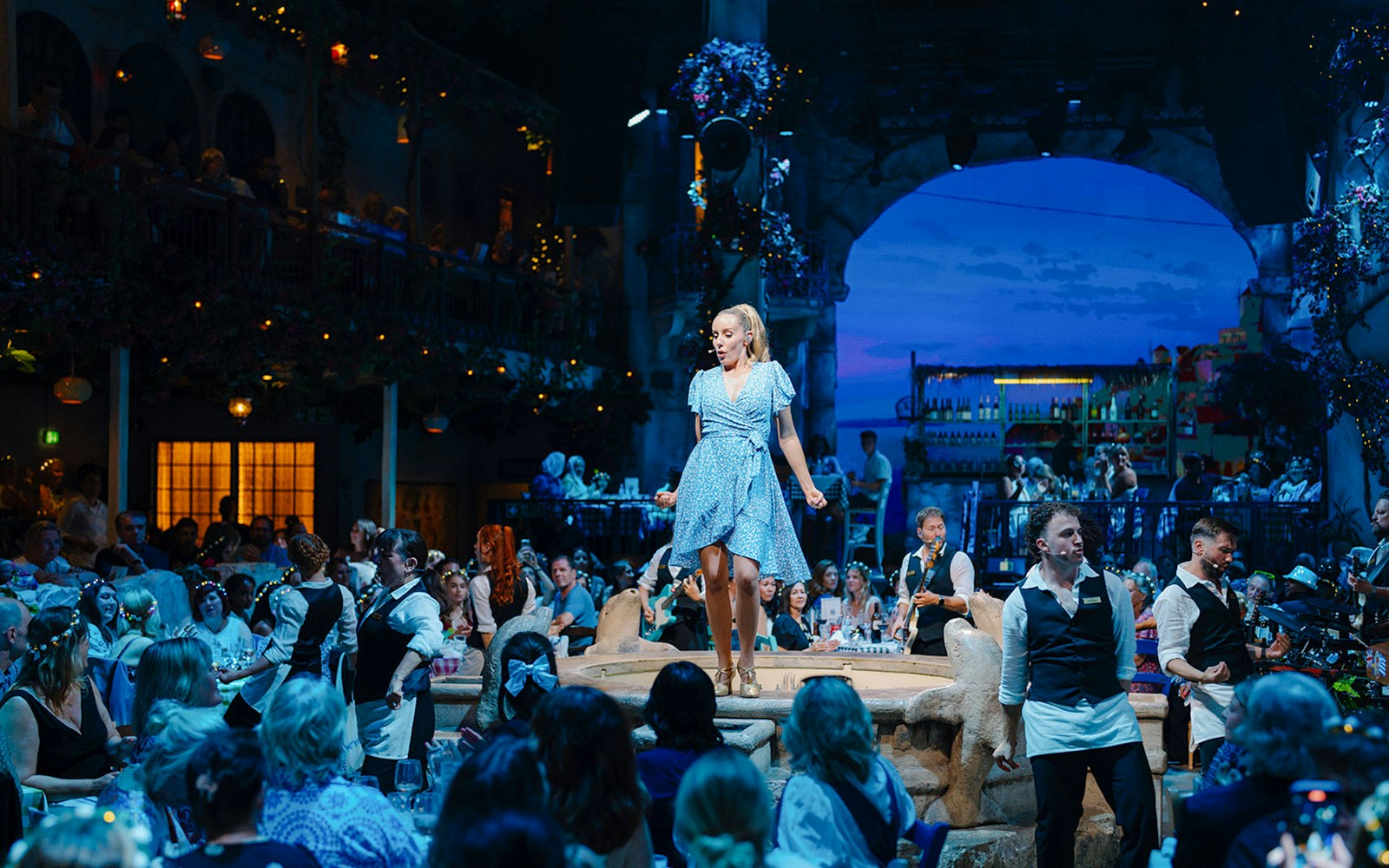 Performer on stage at Mamma Mia! The Party with audience dining in a Greek-themed setting.