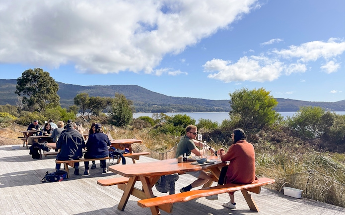 Tour group dining outdoors with scenic Bruny Island coastline in background, Hobart day trip.