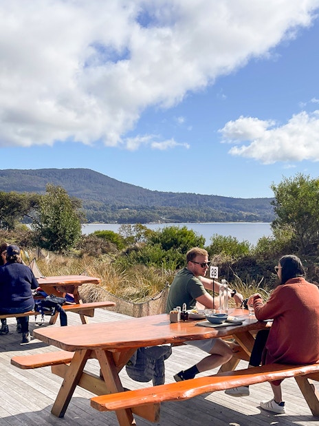 Tour group dining outdoors with scenic Bruny Island coastline in background, Hobart day trip.