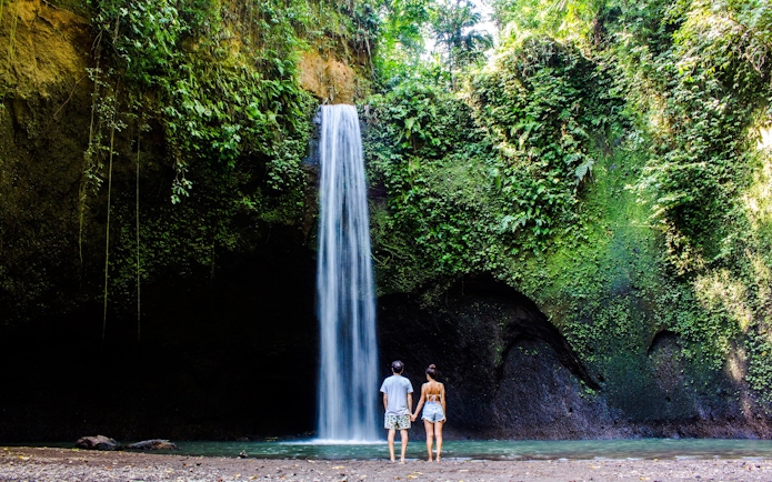 Visitors standing in front of Tibumana Waterfall surrounded by lush greenery in Bali, Indonesia.