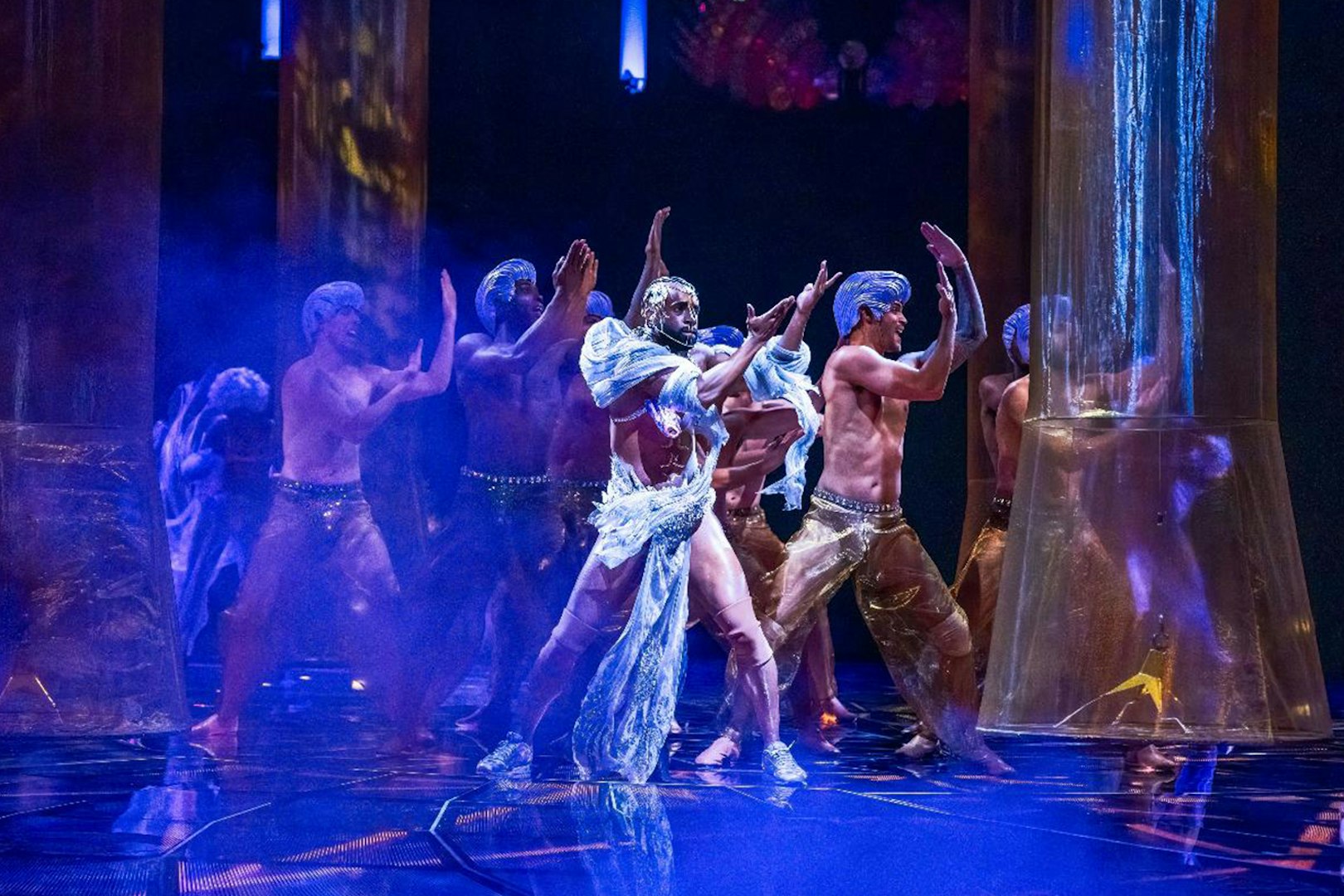 Performers in elaborate costumes dance under blue lighting in a theatrical show.