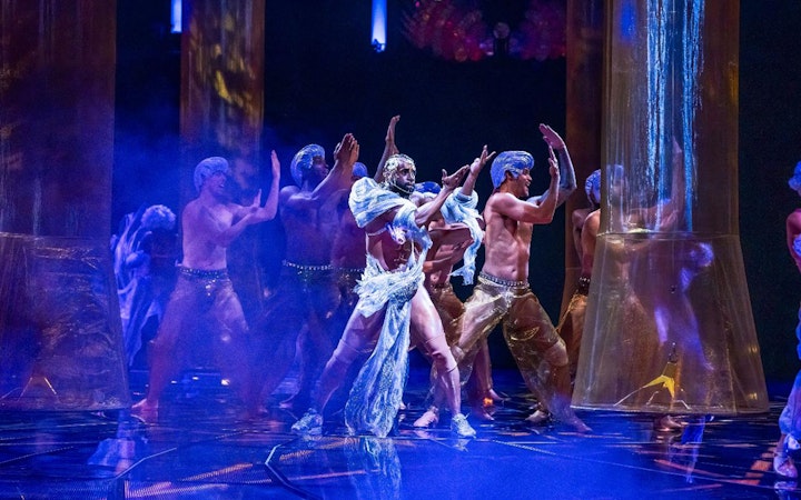 Performers in elaborate costumes dance under blue lighting in a theatrical show.