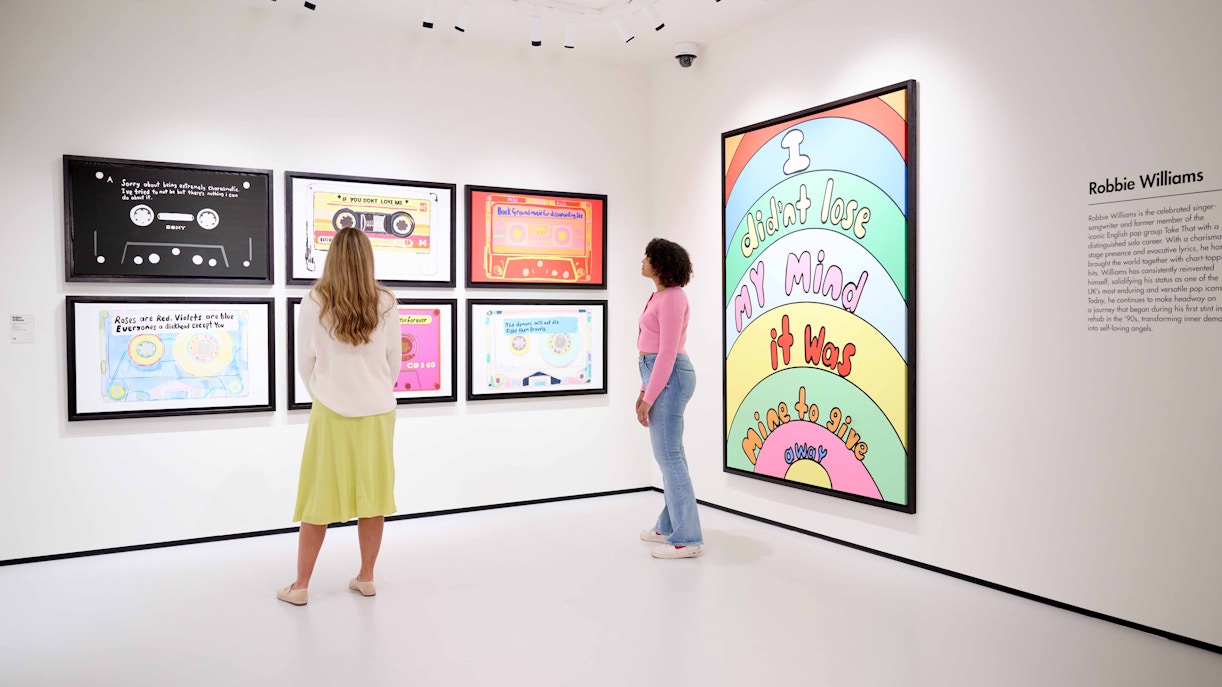 Visitors viewing colorful cassette-themed art in a Robbie Williams exhibition.