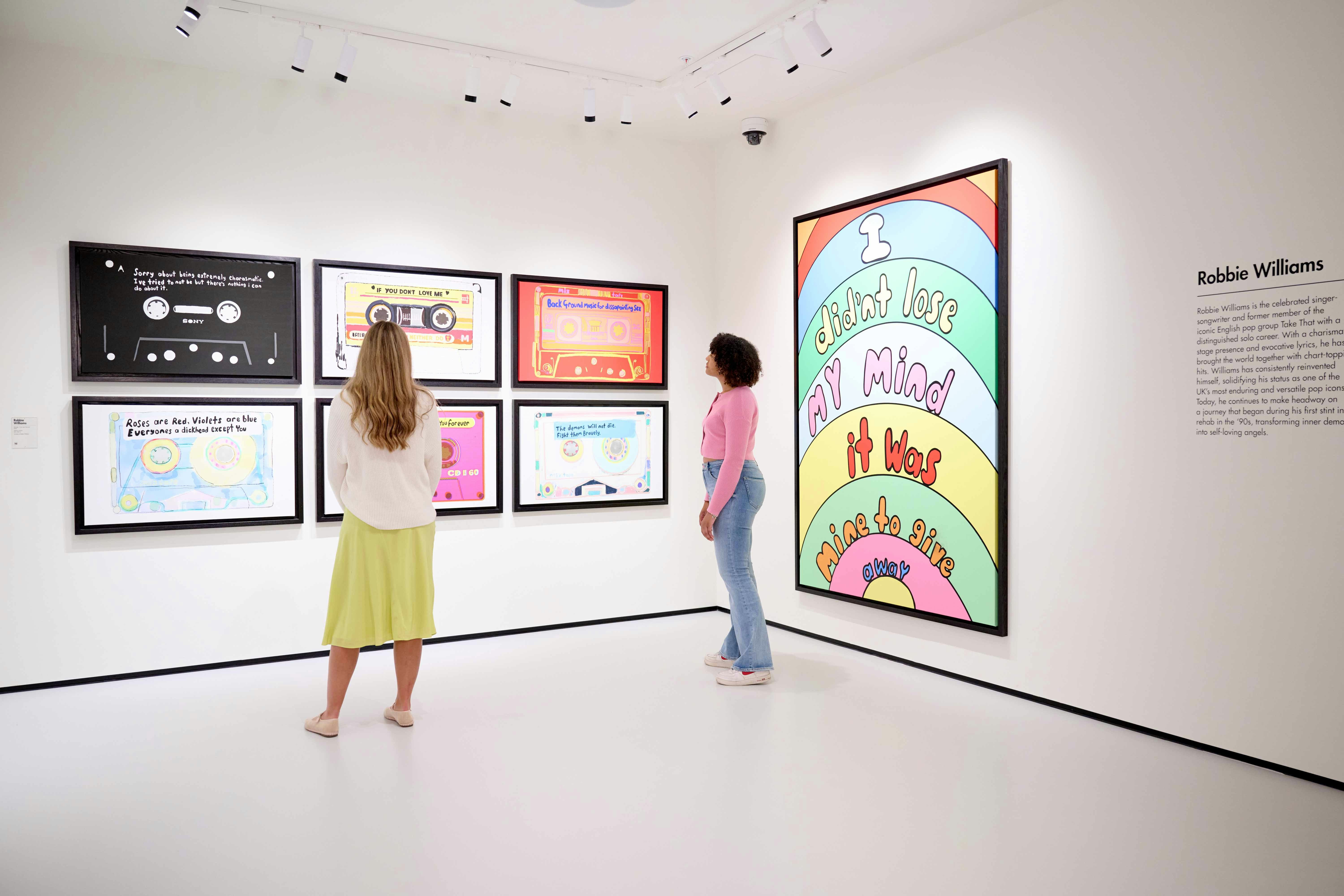 Visitors viewing colorful cassette-themed art in a Robbie Williams exhibition.