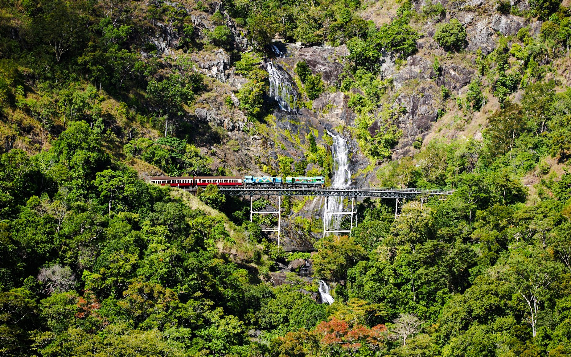 Train crossing a bridge with a waterfall in a lush forest during a scenic tour.