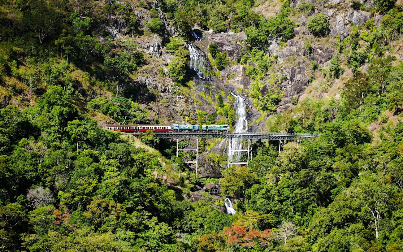 Train crossing a bridge with a waterfall in a lush forest during a scenic tour.