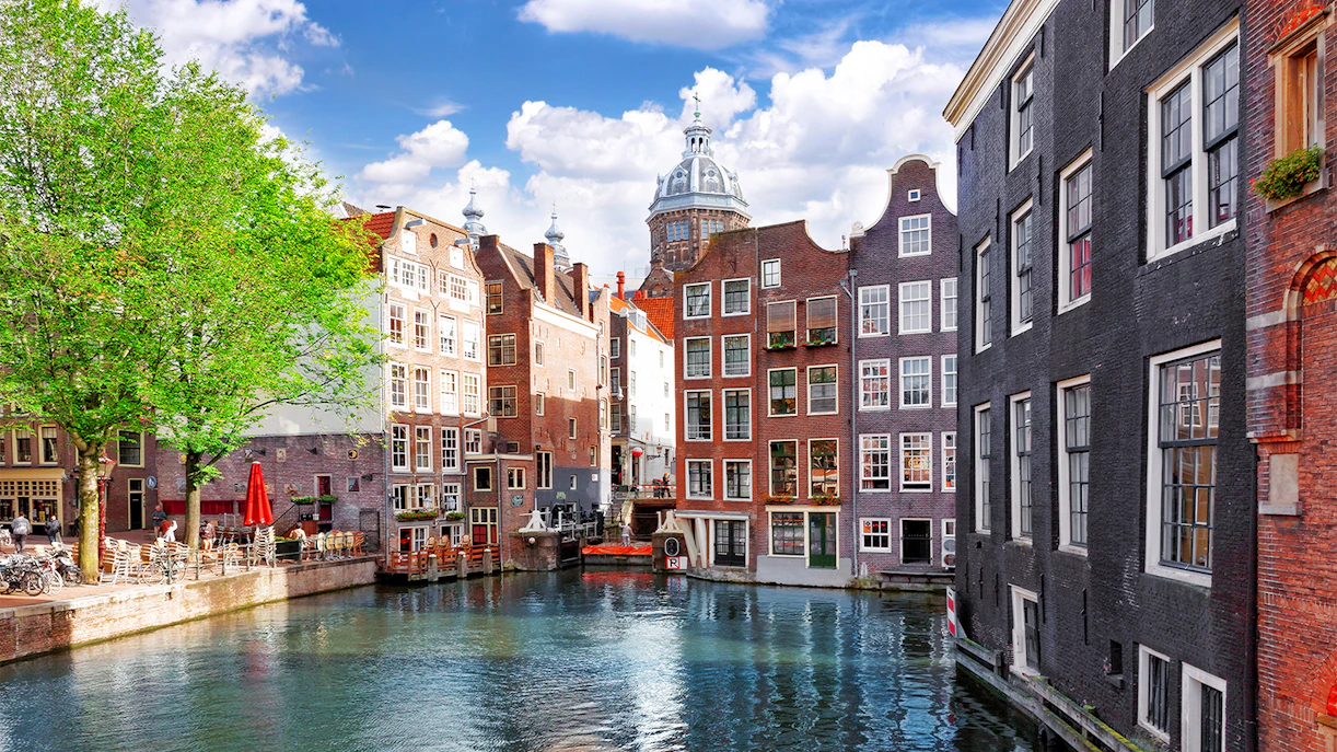 Amsterdam canal with historic buildings and a tree-lined walkway.