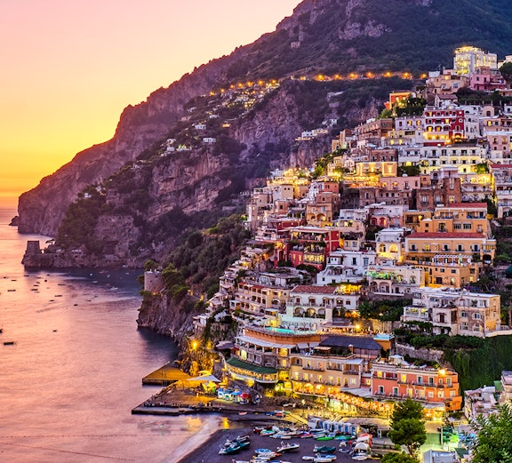 Positano coastline at sunset with colorful hillside buildings and boats on the water.