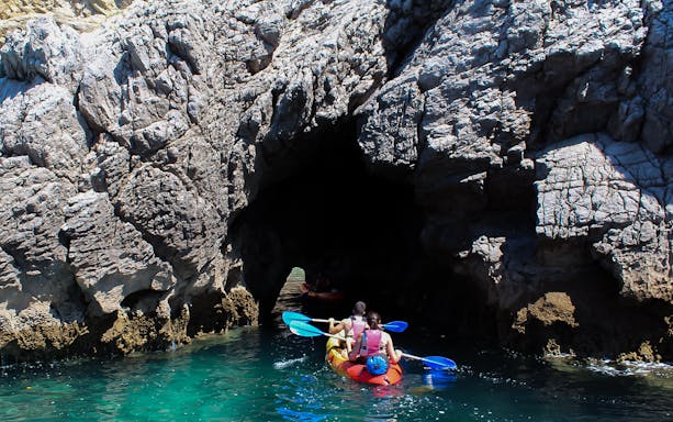 Kayakers paddling in clear waters of Arrábida Natural Park, Lisbon, with rocky cliffs in the background.