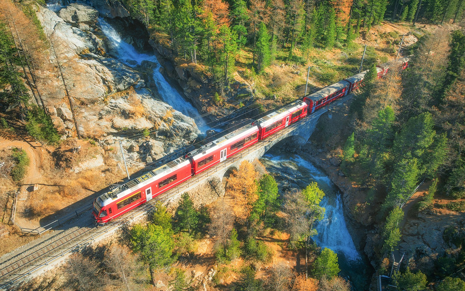 Aerial view of red passenger train traveling across a bridge over a waterfall, surrounded by colorful autumn forest in the swiss alps.