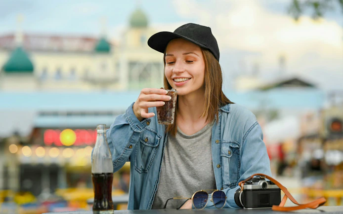 Woman enjoying a cola at an outdoor cafe with a camera and sunglasses on the table.