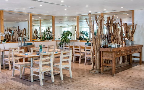 Dining area at Riva Beach Club with wooden tables and chairs, surrounded by plants.