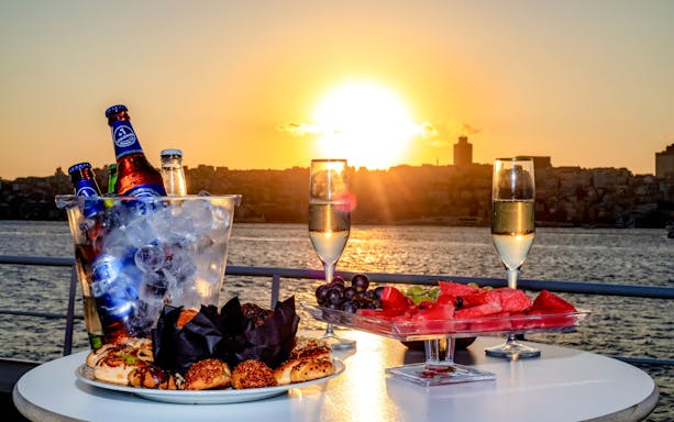 Bosphorus sunset cruise with champagne, fruit platter, and pastries on a table.