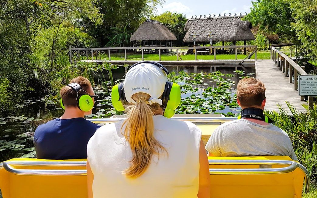Guests on airboat ride in Everglades, viewing lush greenery and thatched huts.