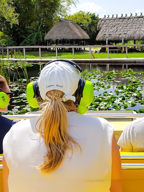Guests on airboat ride in Everglades, viewing lush greenery and thatched huts.